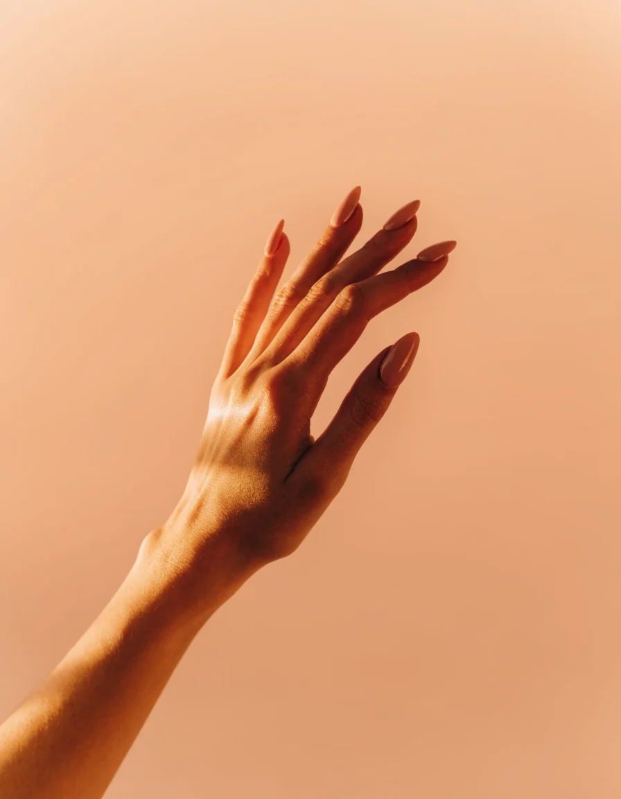 A raised hand and forearm with warm nude almond nails against a soft peach backdrop